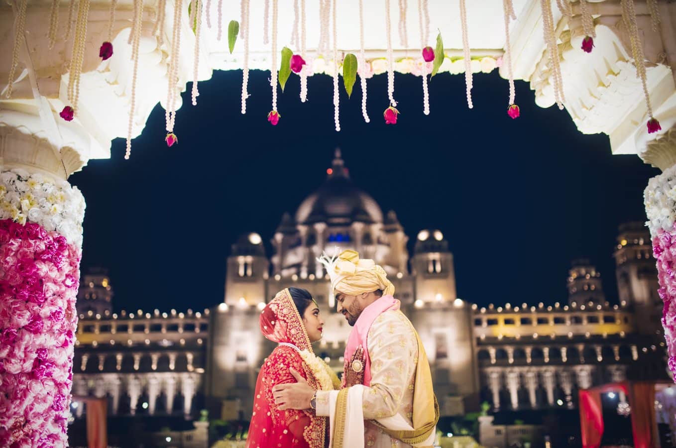 Elegant traditional Indian wedding couples at night in front of a grand historic building with floral decor.